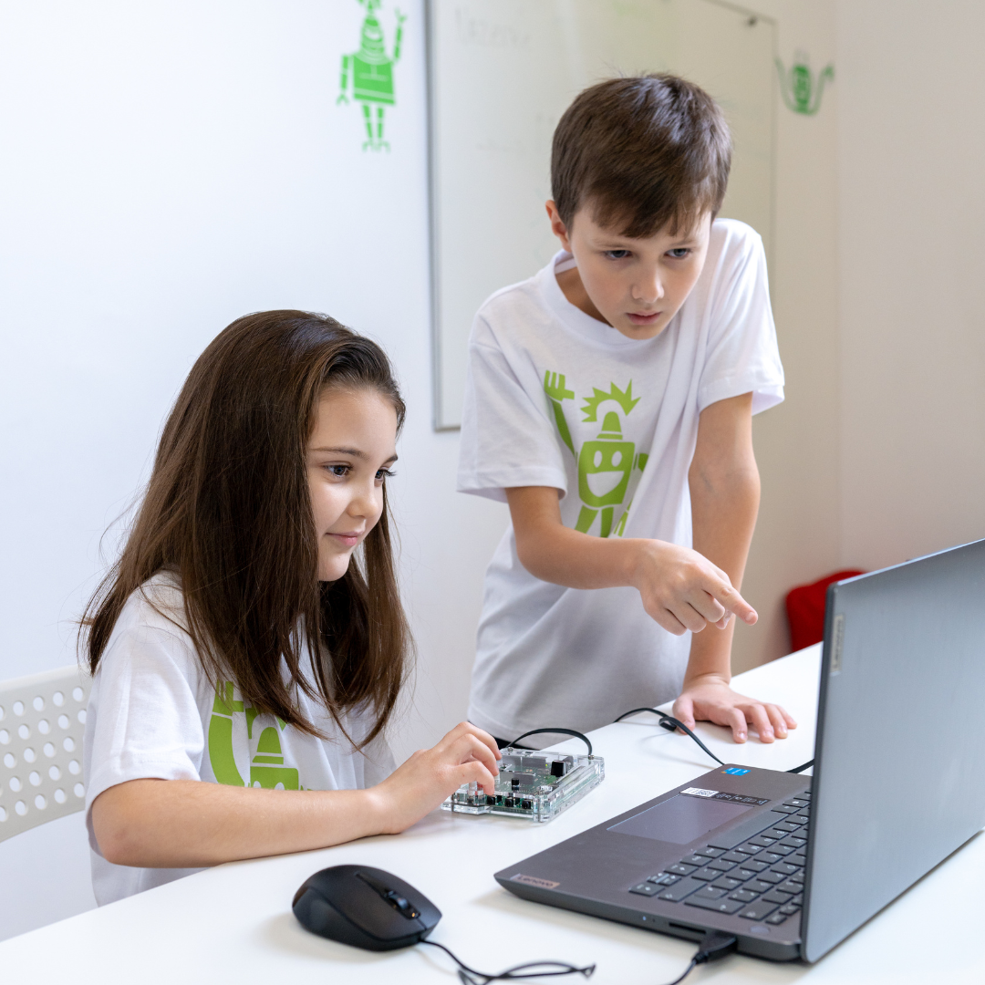 Zwei Kinder in weißen Robbo-Club-T-Shirts arbeiten gemeinsam an einem Elektronikprojekt mit einem Laptop und einer Mikrocontroller-Platine.