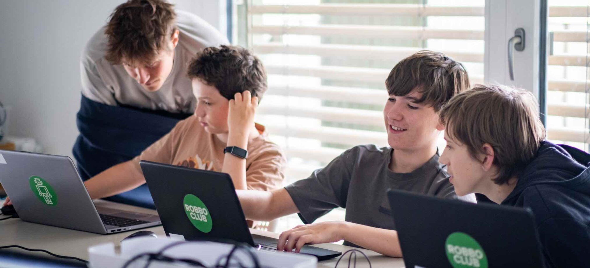 Jugendliche ab 13 Jahren arbeiten gemeinsam an Laptops im Robbo Club Feriencamp und unterstützen sich beim Programmieren.