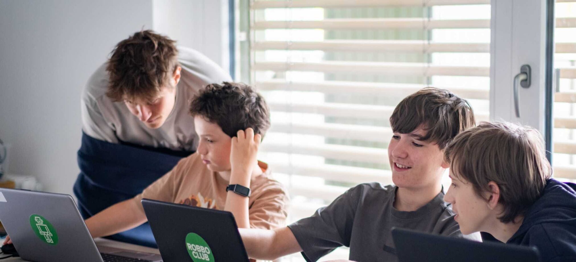 Vier Jugendliche arbeiten im Robbo Club gemeinsam an Laptops, während ein Betreuer ihnen beim Programmieren hilft.