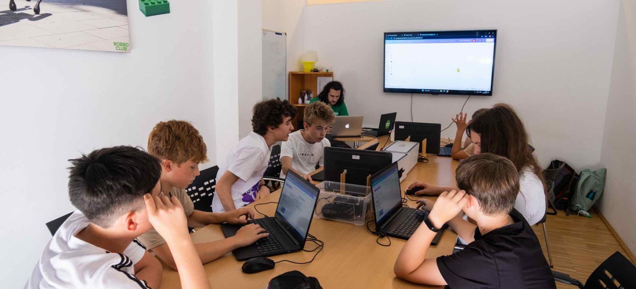 Jugendliche ab 15 Jahren arbeiten im Robbo Club Feriencamp konzentriert an Laptops, während der Trainer vorne am Bildschirm erklärt.