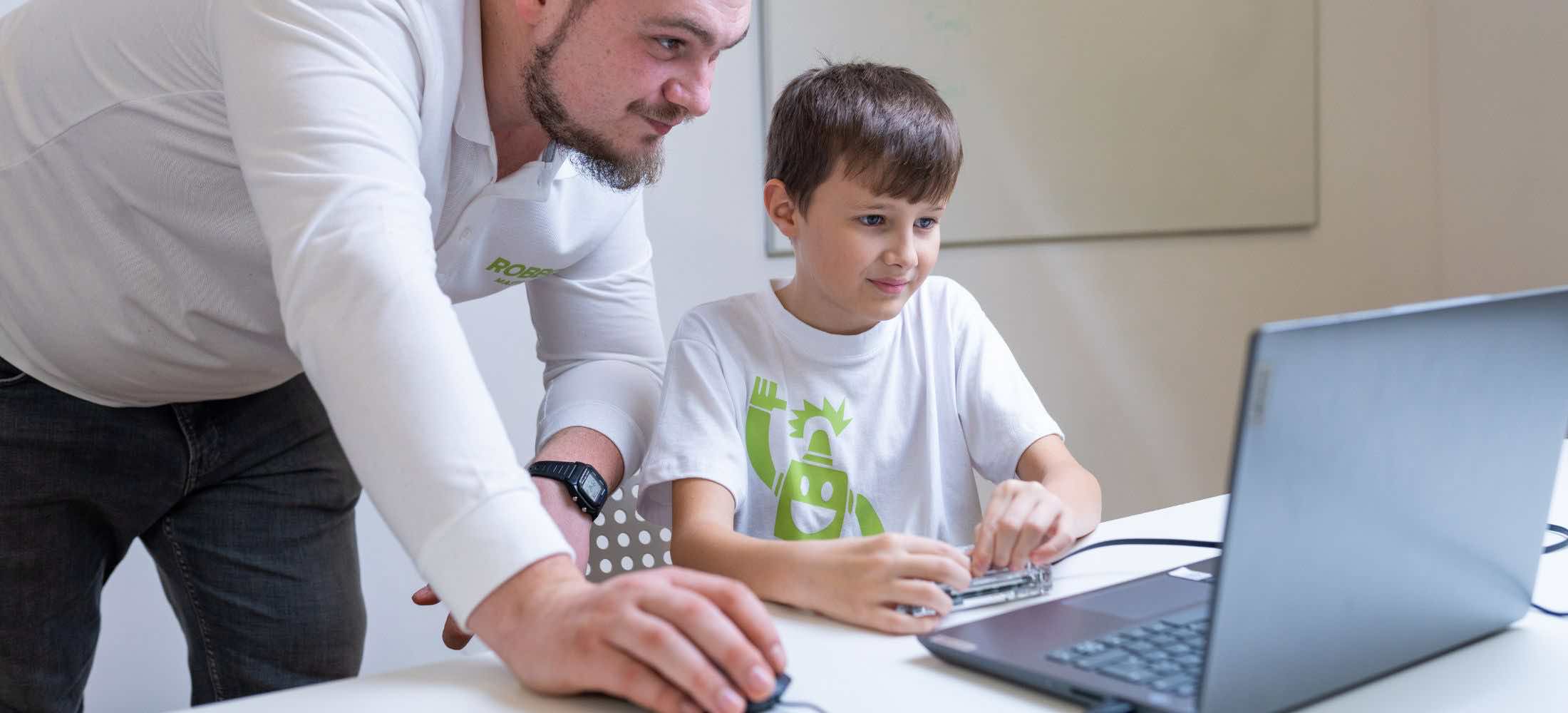 Kind arbeitet im Robbo Club mit einer Elektronikplatine am Laptop, unterstützt von einem Trainer.