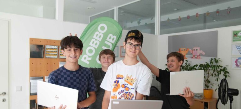 Vier Jugendliche im Robbo Club posieren fröhlich mit ihren Laptops vor einem grünen Robbo Club Banner.