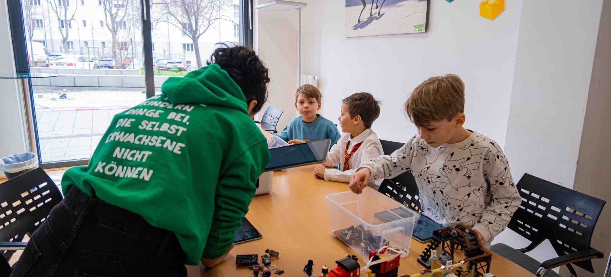 Kinder sitzen an einem Tisch im Robbo Club und bauen LEGO-Modelle. Ein Trainer unterstützt die Gruppe, während die Kinder konzentriert Bauteile sortieren und mit Tablets arbeiten.