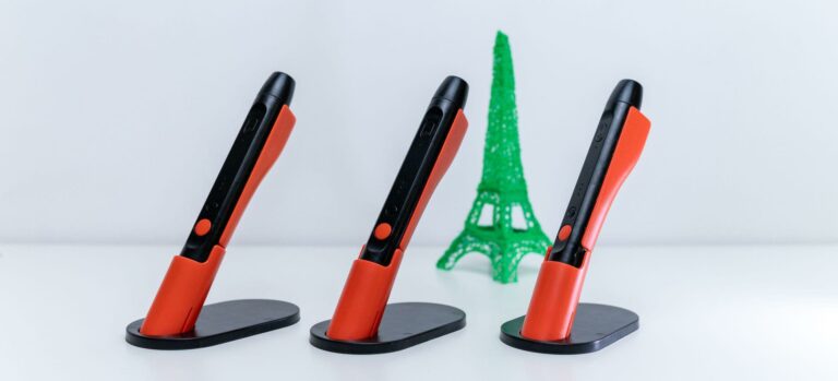 Drei orange-schwarze 3D-Stifte stehen in Halterungen, im Hintergrund ist ein grünes 3D-Modell des Eiffelturms zu sehen.