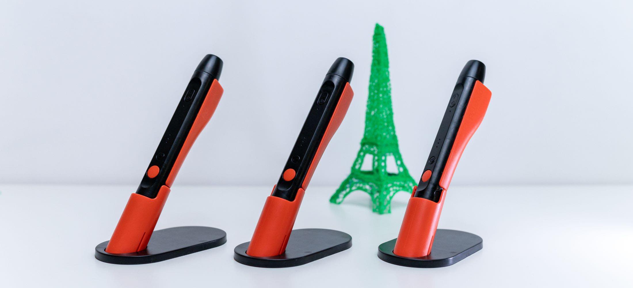 Drei orange-schwarze 3D-Stifte stehen in Halterungen, im Hintergrund ist ein grünes 3D-Modell des Eiffelturms zu sehen.