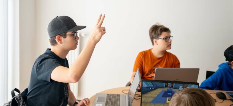 Jugendliche im Robbo-Club-Kurs arbeiten an Laptops, ein Junge hebt die Hand, um eine Frage zu stellen.