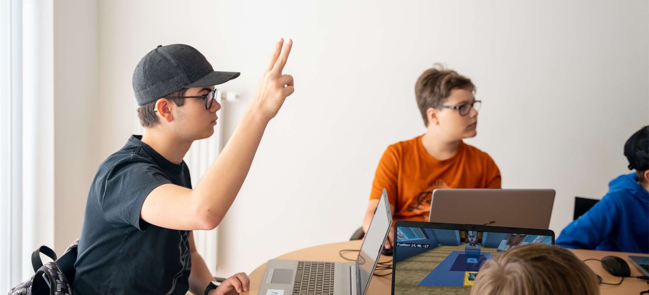 Jugendliche im Robbo-Club-Kurs arbeiten an Laptops, ein Junge hebt die Hand, um eine Frage zu stellen.