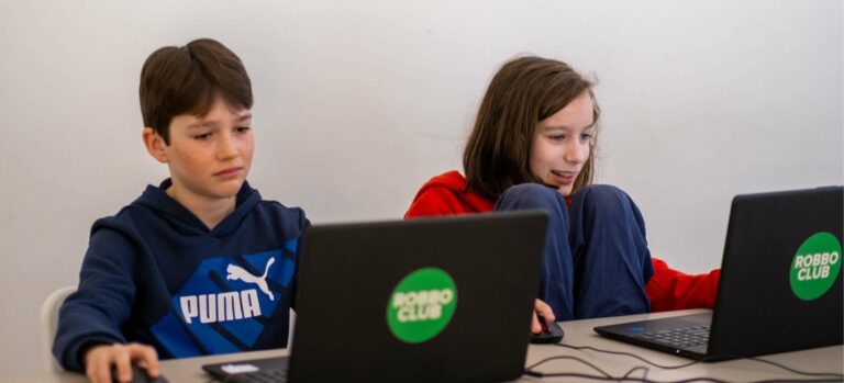 Zwei Jugendliche sitzen am Laptop im Robbo Club und arbeiten an einem Computerprojekt; einer wirkt konzentriert, der andere lächelt motiviert.