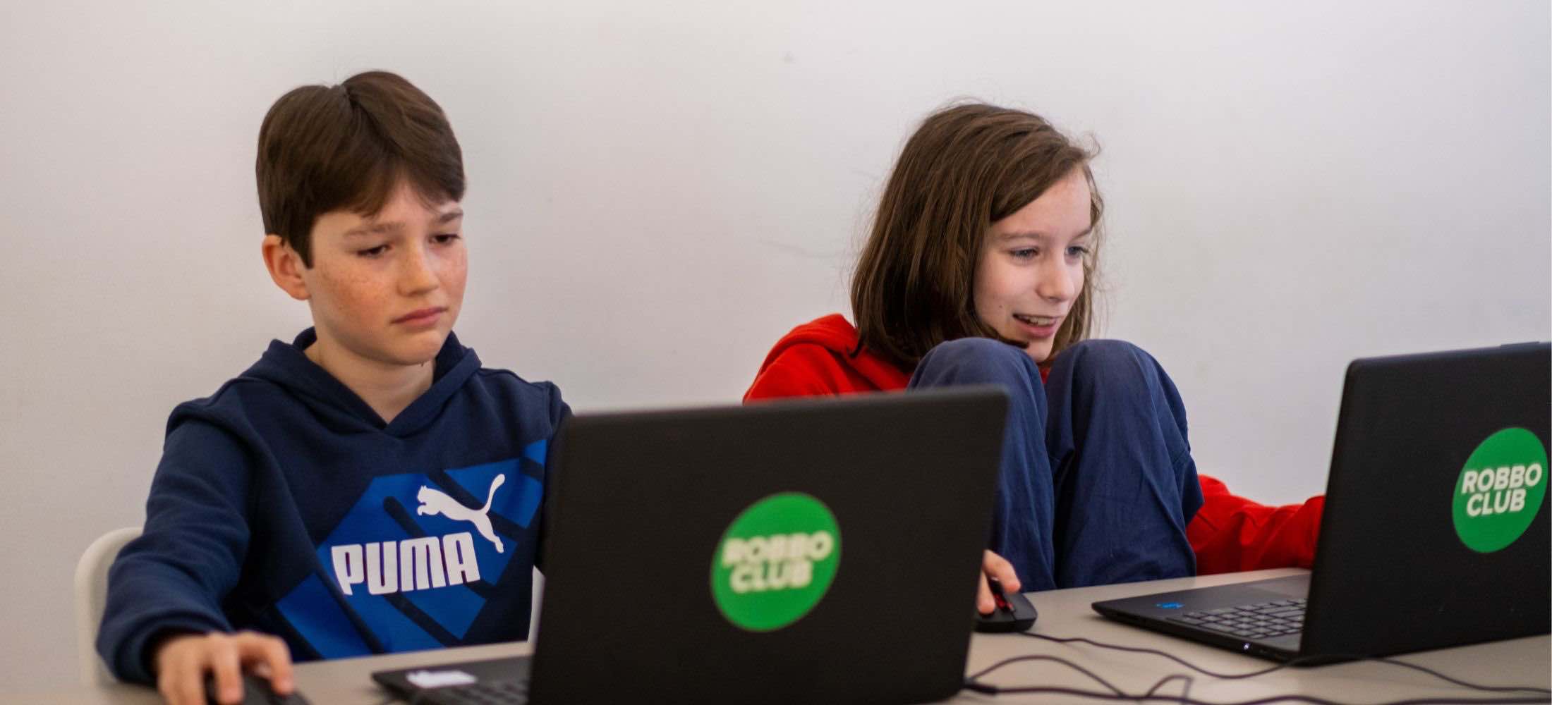 Zwei Jugendliche sitzen am Laptop im Robbo Club und arbeiten an einem Computerprojekt; einer wirkt konzentriert, der andere lächelt motiviert.