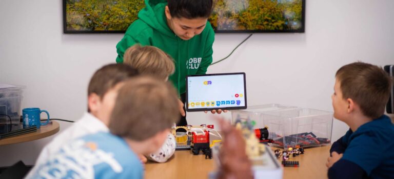 Eine Trainerin zeigt Kindern auf einem Tablet ein LEGO-Robotik-Programmierinterface, während LEGO-Modelle auf dem Tisch stehen.