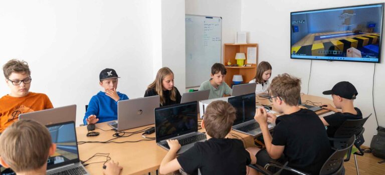 Kinder sitzen mit Laptops im Unterricht und programmieren Minecraft-Welten, während eine große Bildschirmanzeige das Spiel zeigt.