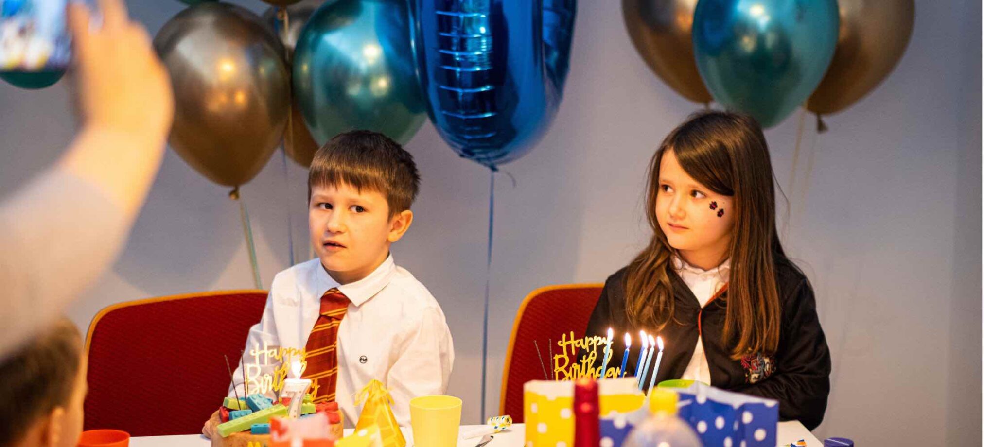 Kinder sitzen bei einem Indoor-Kindergeburtstag im Robbo Club am festlich dekorierten Tisch mit Luftballons und Geburtstagskuchen.