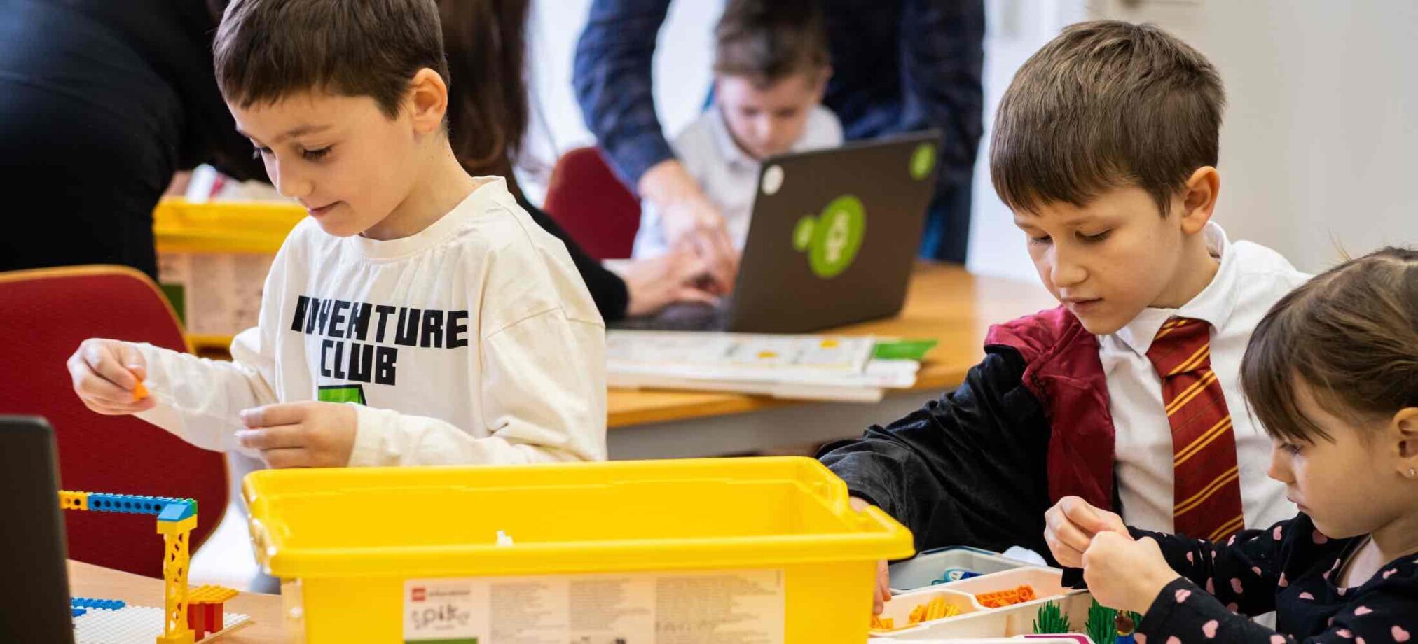 Kleine Kinder bauen mit LEGO-Elementen und sitzen gemeinsam am Tisch, während im Hintergrund Erwachsene unterstützen. Die Kinder tragen bunte Kleidung und arbeiten konzentriert.
