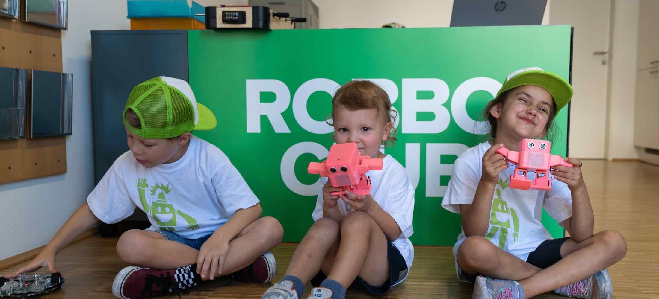 Drei Kinder im Alter von etwa sechs Jahren sitzen im Robbo Club und halten stolz ihre selbstgebauten Roboter.