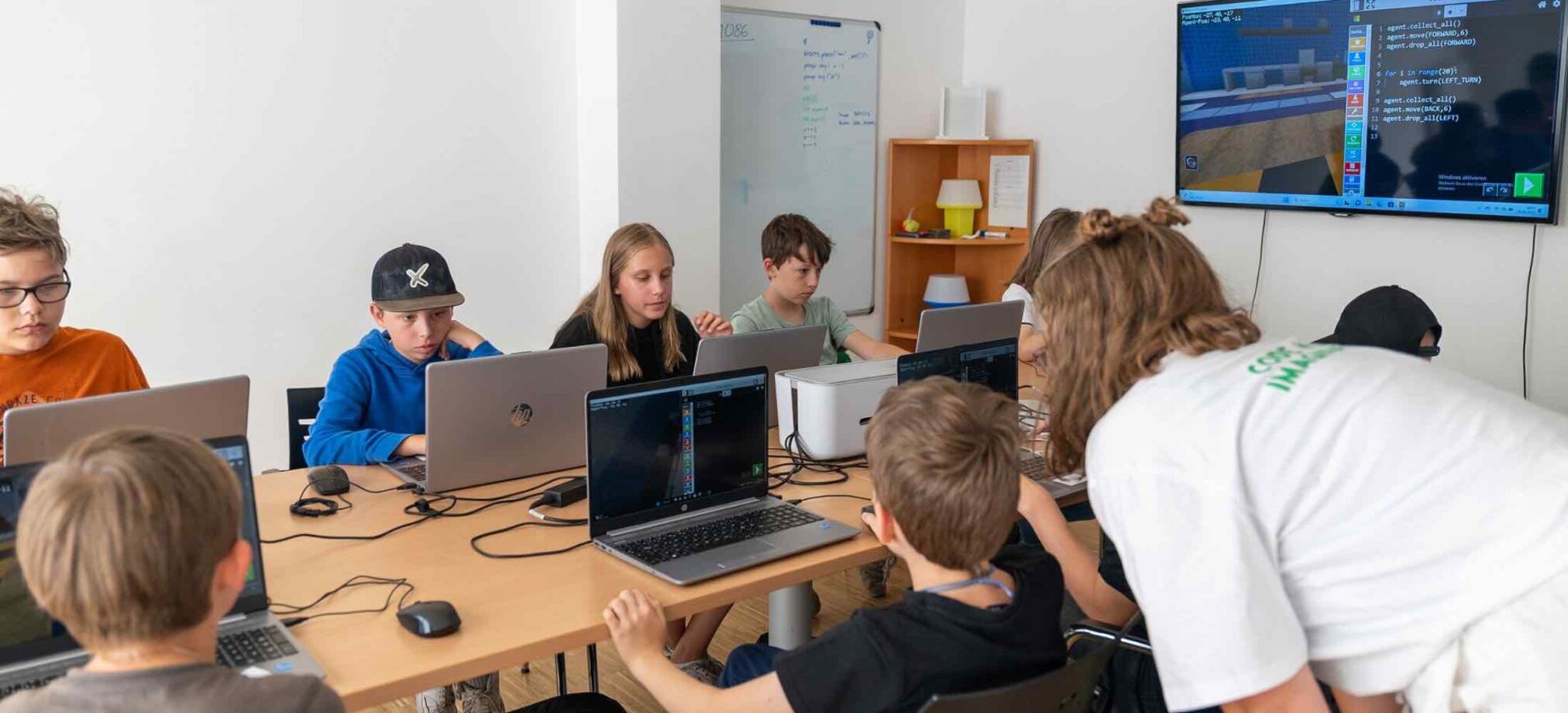 Kinder programmieren im IT-Feriencamp des Robbo Club an Laptops, während eine Trainerin sie individuell unterstützt.