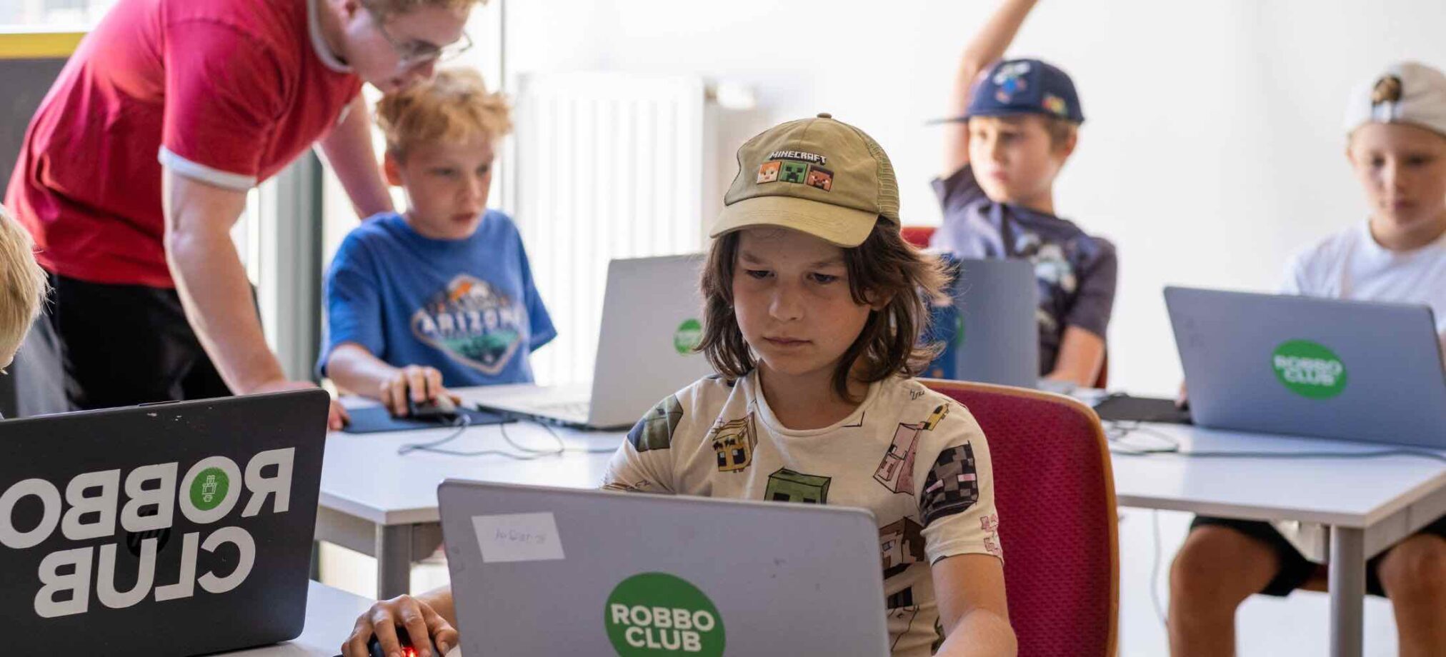 Kinder arbeiten im Computer-Feriencamp des Robbo Club konzentriert an Laptops, unterstützt von einem Trainer.