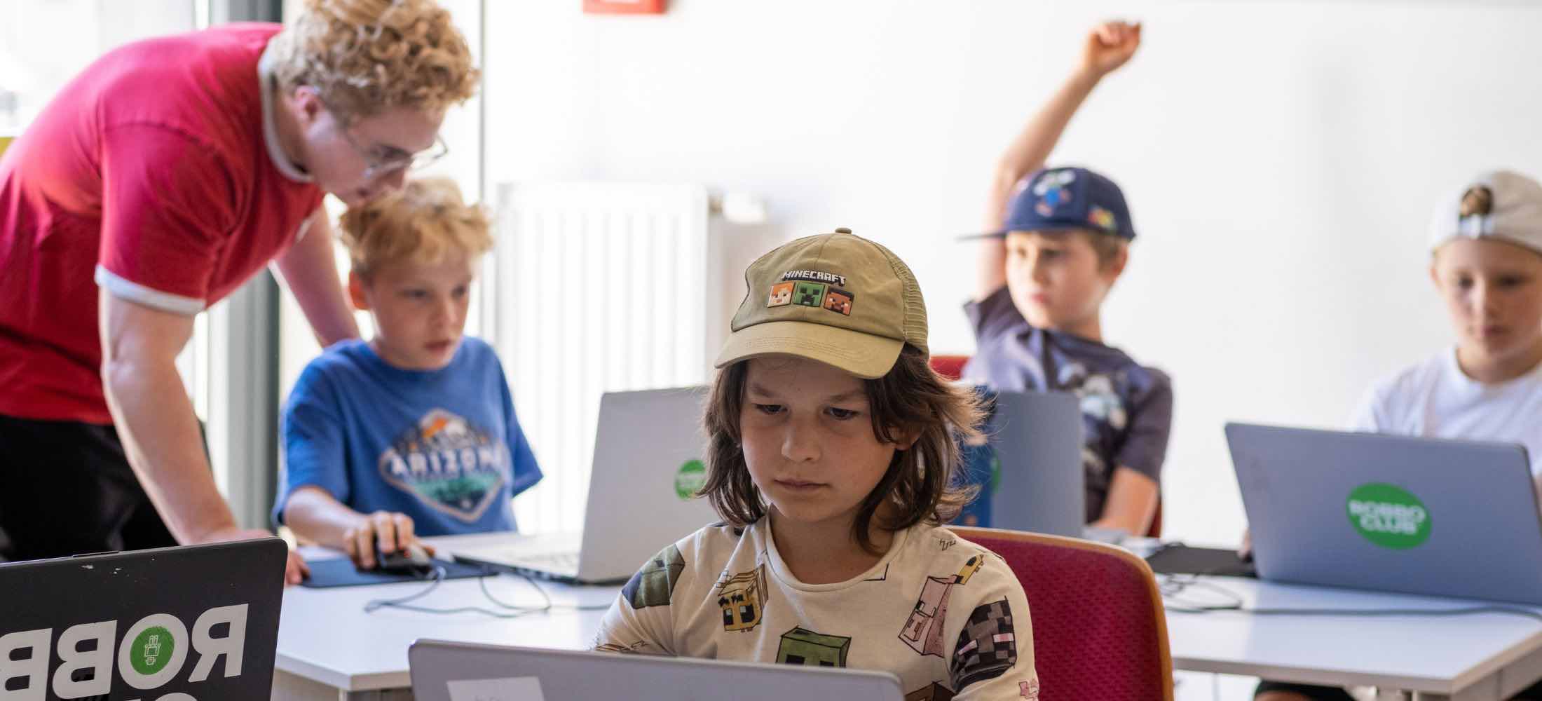 Kinder programmieren und spielen im Minecraft-Feriencamp des Robbo Club an Laptops, unterstützt von einem Trainer.