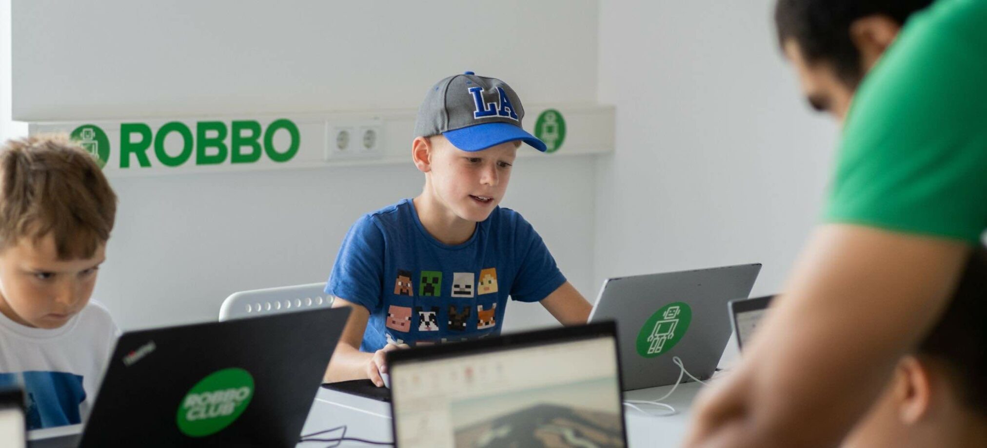 Kinder programmieren im Robbo Club Feriencamp an Laptops und werden von einem Trainer begleitet.