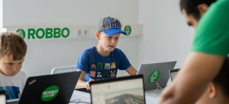 Kinder programmieren im Robbo Club Feriencamp an Laptops und werden von einem Trainer begleitet.