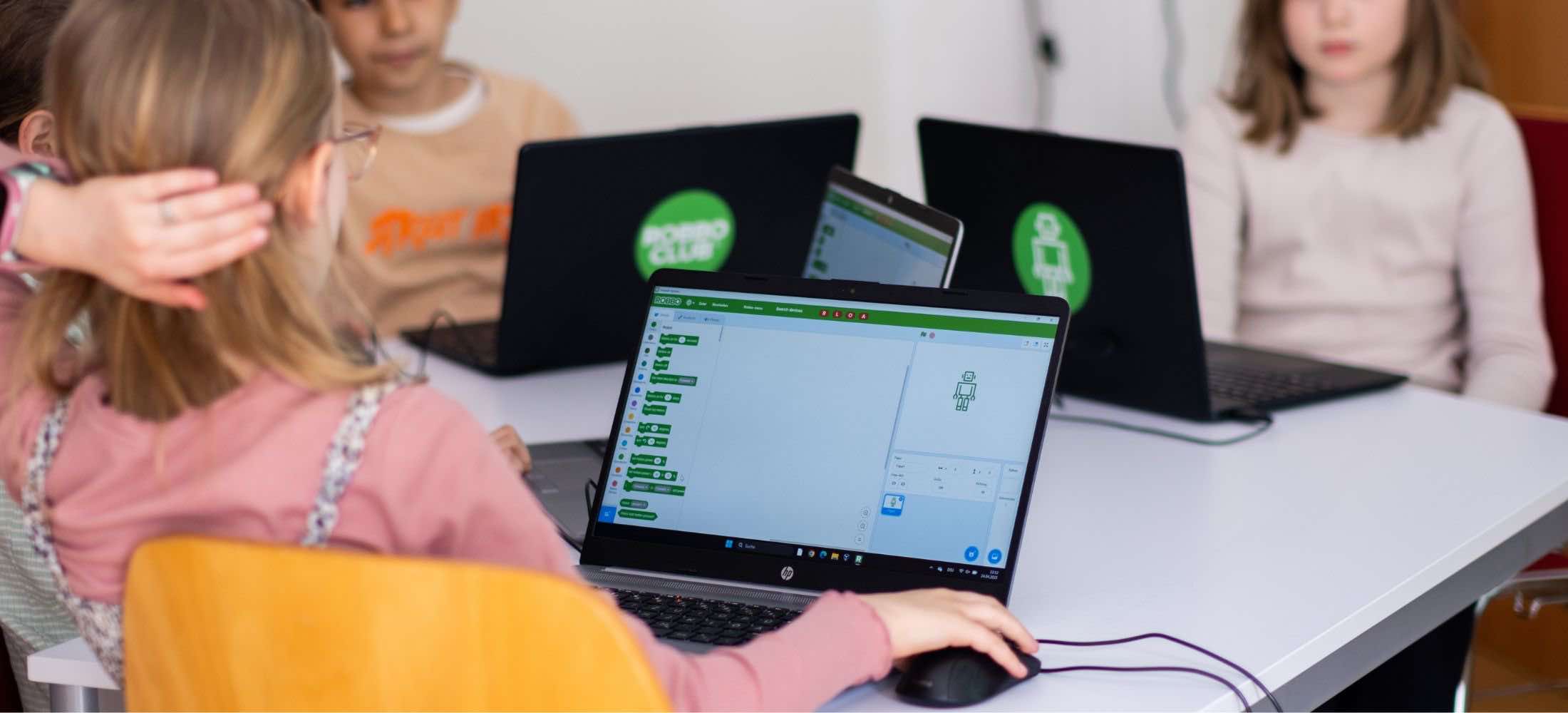 Kinder sitzen in einem Programmier-Workshop des Robbo Clubs und arbeiten mit visuellen Coding-Blöcken am Laptop.