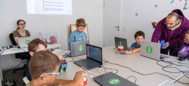 Kinder programmieren im Robbo Feriencamp an Laptops und steuern kleine Lernroboter, unterstützt von zwei Trainern.