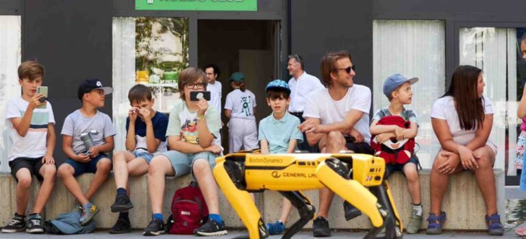 Kinder des Robbo Club Sommercamps beobachten begeistert einen laufenden Roboterhund von Boston Dynamics vor dem Clubgebäude.