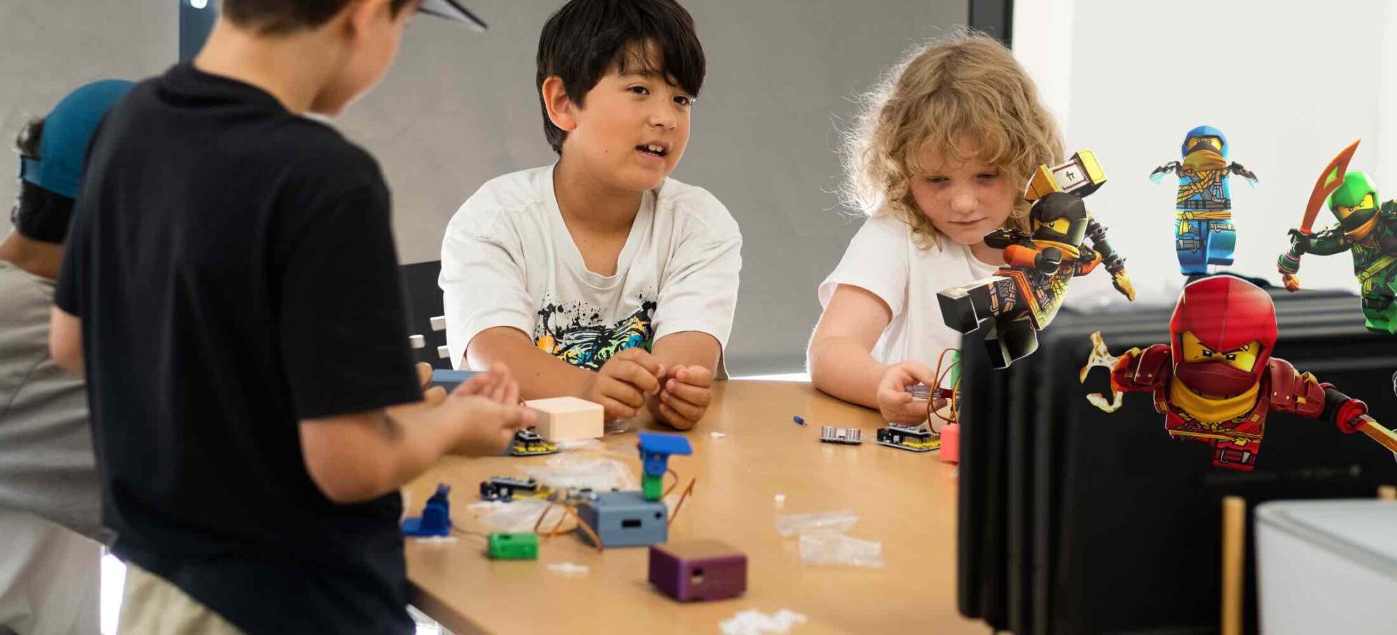 Kinder arbeiten im Robbo Club an kleinen Robotikprojekten, während Ninja-Figuren als thematische Dekoration den Tag-des-Ninja-Workshop begleiten.