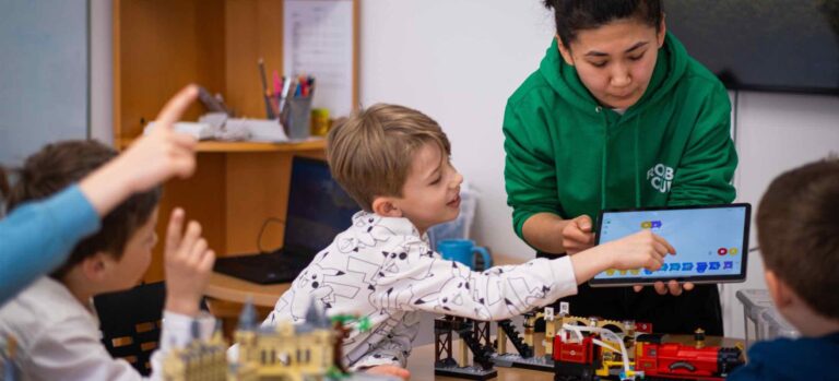Kinder programmieren im Robbo Club Weihnachtsferiencamp mithilfe eines Tablets und steuern LEGO-Modelle, unterstützt von einer Trainerin.