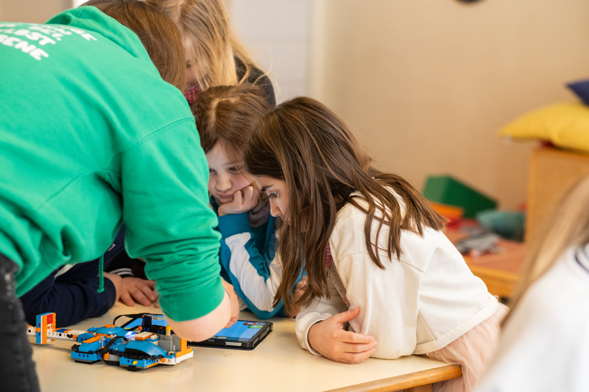 ROBBO Trainerin im grünen Hoodie hilft zwei Mädchen beim Programmieren eines LEGO-Roboters am Tablet