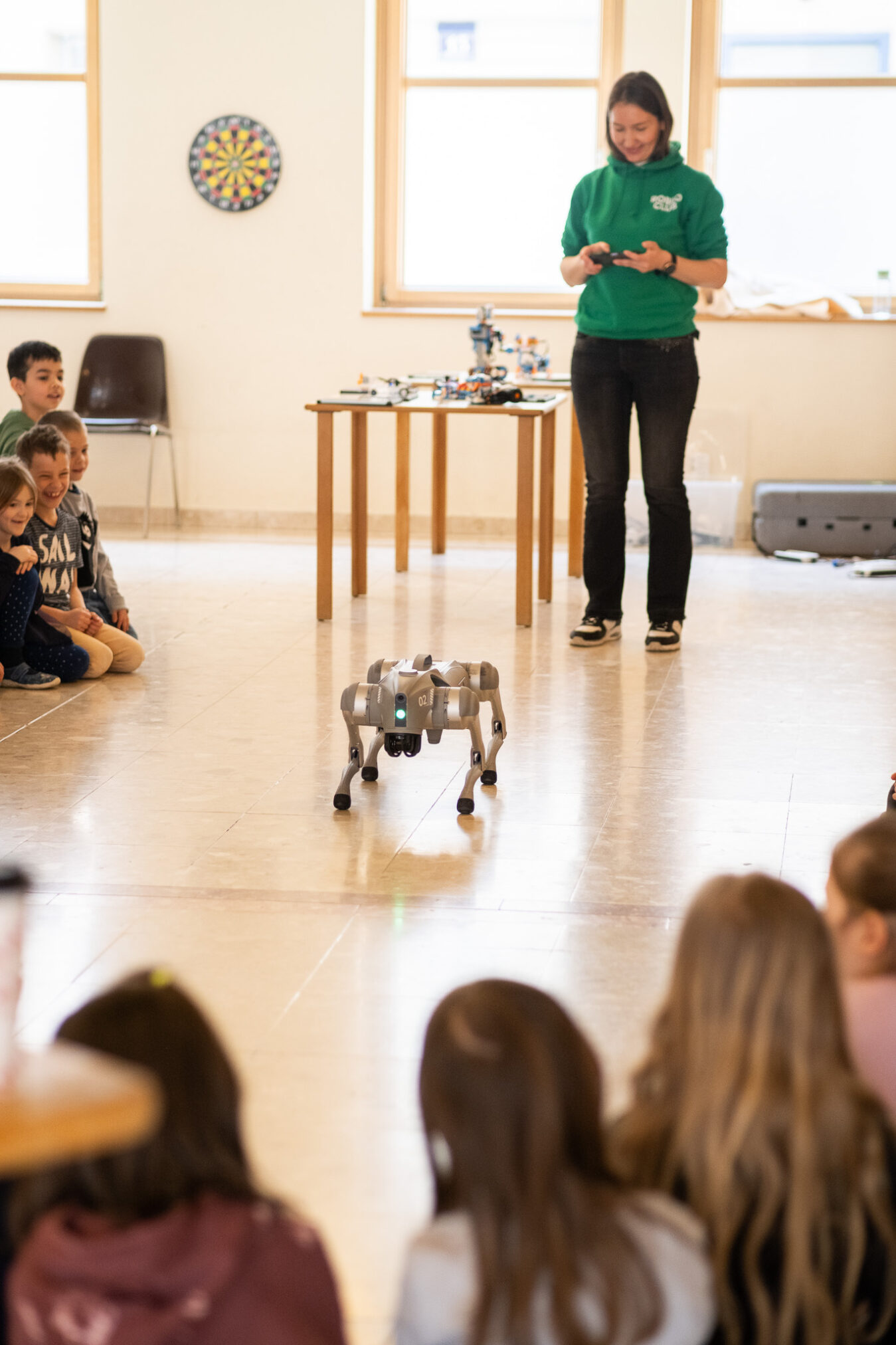 ROBBO Trainerin im grünen Hoodie steuert einen Roboterhund vor einer Schulklasse, die Kinder sitzen im Halbkreis am Boden